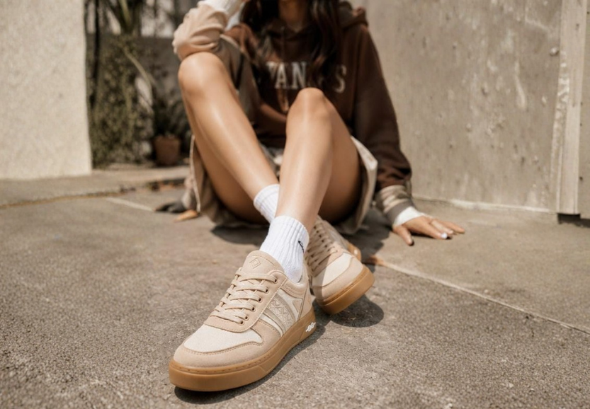 Person seated on a pavement in a light outfit, wearing cream NAAW sneakers with white socks